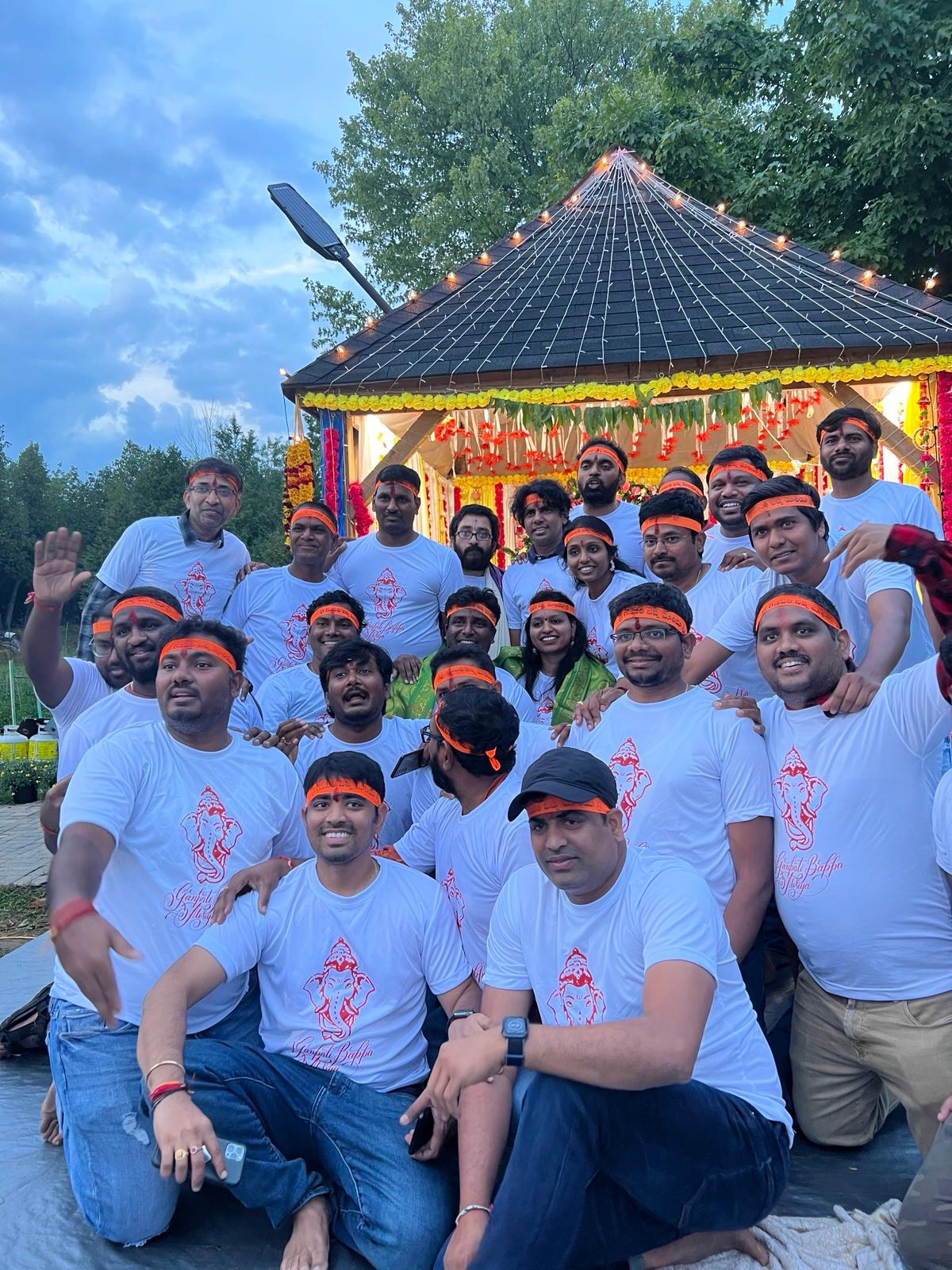 Community members wearing matching Ganesh festival t-shirts in front of a decorated outdoor mandap, 2022