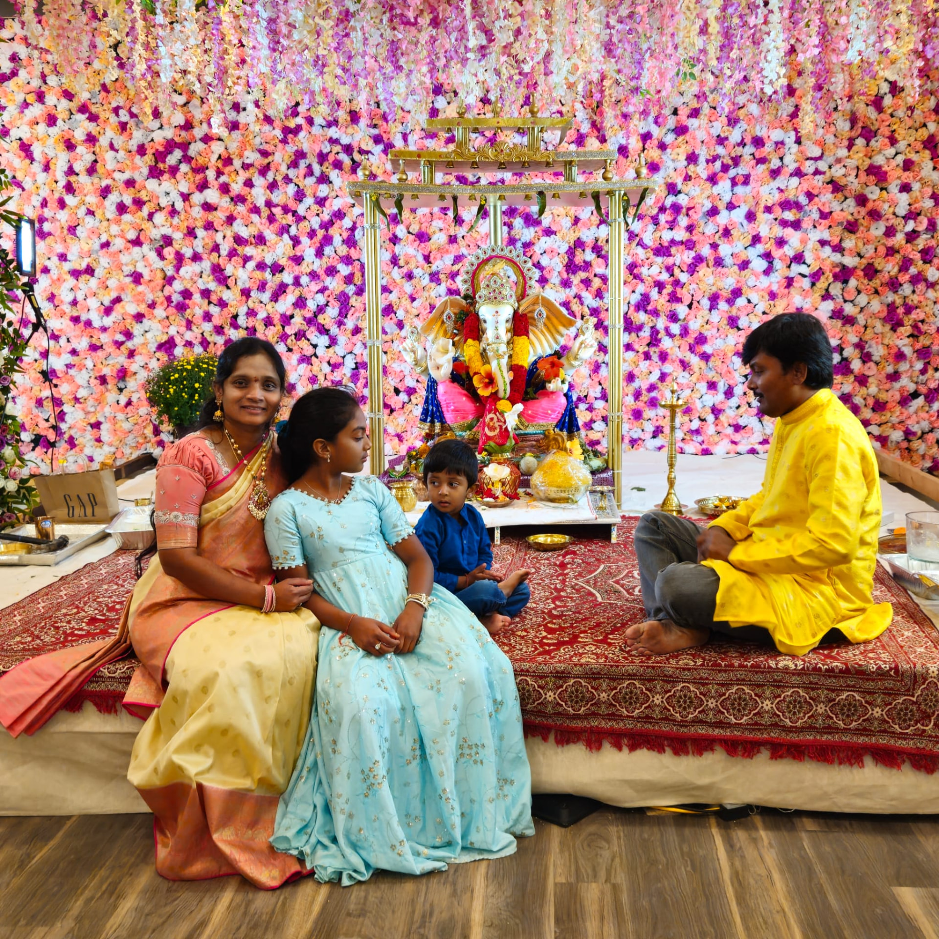 A family seated before an elaborate pink and purple floral Ganesh mandap, 2024